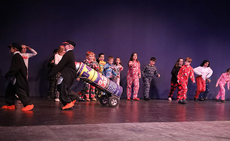 Children dressed in pajamas watching another group of children in penguin costumes carrying a giant can of WD-40 on a dolly