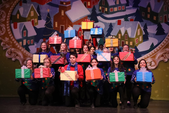 People holding brightly wrapped gifts in front of a Dickens village backdrop.
