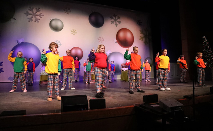 Children dressed in plaid pants and bright shirts dance in front of an ornament drop.