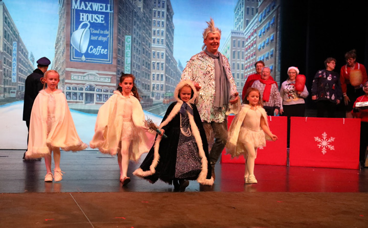 A tall man wearing sparkling clothing walks with children dressed as snow angels against a backdrop of a New York city scene.