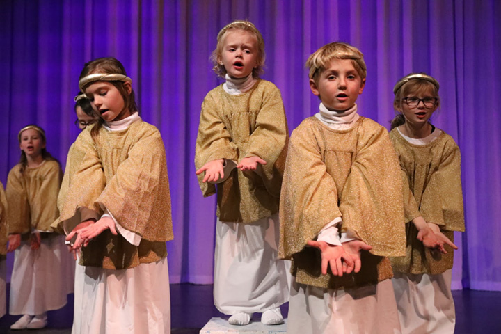 Young children dressed in white gowns, sparkly gold capes and halos sing.
