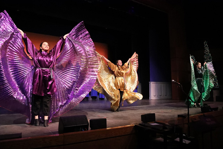 Three young women dance wearing outstretched colored isis wings.