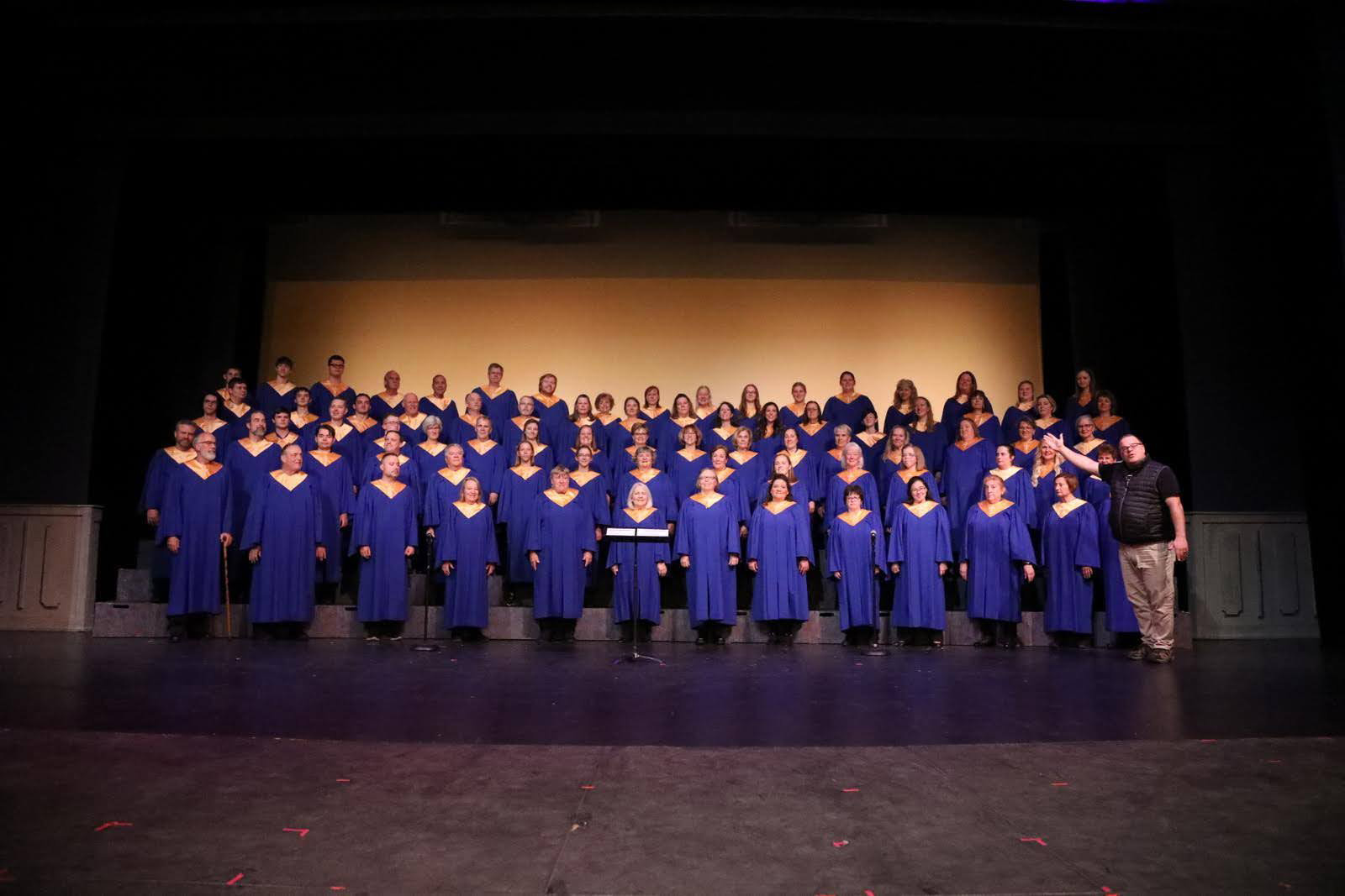 Adults dressed in blue choir robes and gold stoles on stage with director.