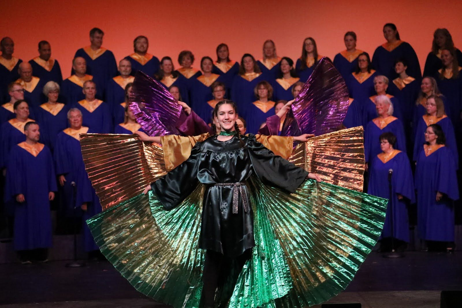 Three dancers in satin outfits with matching isis wings pose in front of a choir.