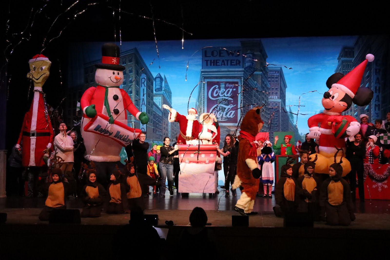 A Christmas parade scene featuring Santa and Mrs. Claus in a sleigh surrounded by floats against a New York Times Square drop.