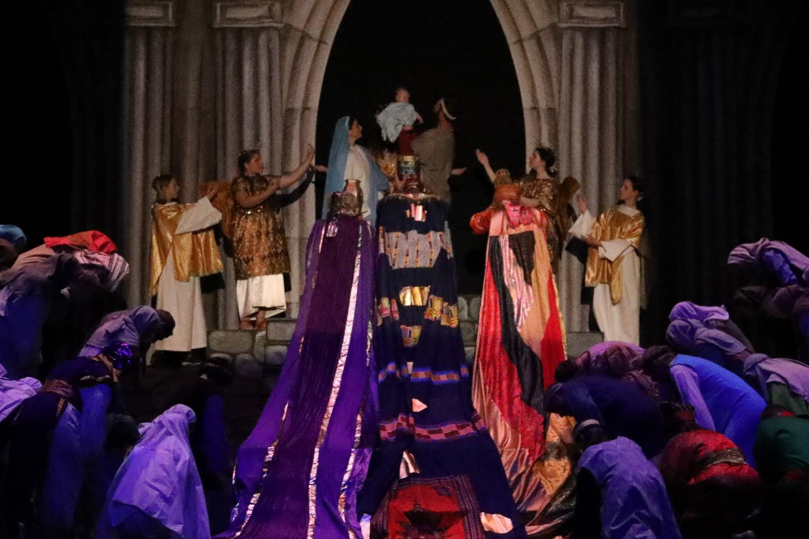 Joseph holds baby Jesus, Mary stands next to them with angels on either side.  Three kings in sumptous costumes and their entourages kneel before them.